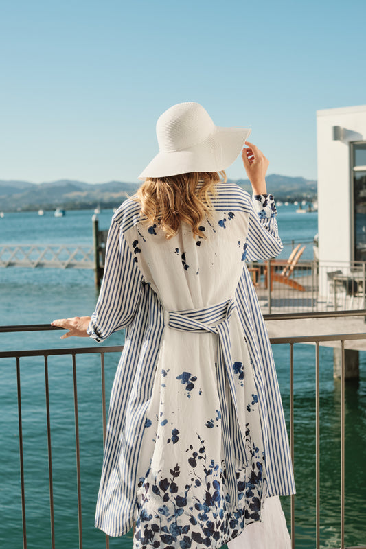 Woman in a floral dress and wide-brimmed hat standing by a waterfront with mountains in the background.