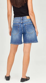 Rear view of a model wearing blue denim shorts with a black top and black flats, shown against a light gray studio background.
