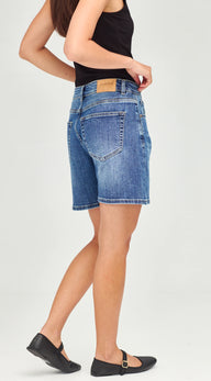 A model wearing blue denim shorts with a black tank top and black flats, shown from a side-back angle against a light gray studio background.