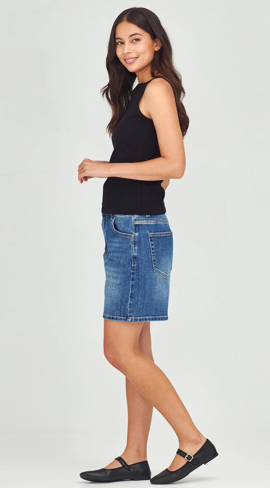 A woman modeling a blue denim mini skirt paired with a black sleeveless top and black Mary Jane flats, standing against a light gray studio background.