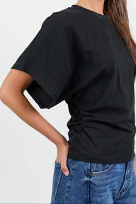 A close-up back view of a woman wearing a black short-sleeve t-shirt with ruching at the side and blue jeans, against a plain light gray background.