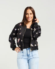 Woman wearing a black bomber jacket with floral patterns, black top, and light blue jeans on a white background