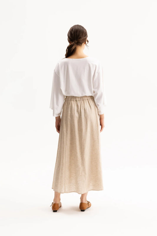 A woman shown from behind wearing a white long-sleeve top and a beige midi skirt, standing against a plain white studio background.