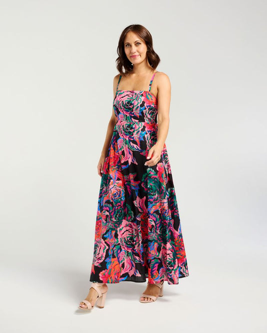 A woman modeling a sleeveless maxi dress with a vibrant floral print in pink, blue, green, and orange on a black background, paired with beige heeled sandals, against a light gray studio backdrop.