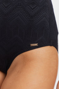 Close-up of a person wearing black textured swimwear bottoms with a geometric pattern and a small gold-tone brand tag near the hem.