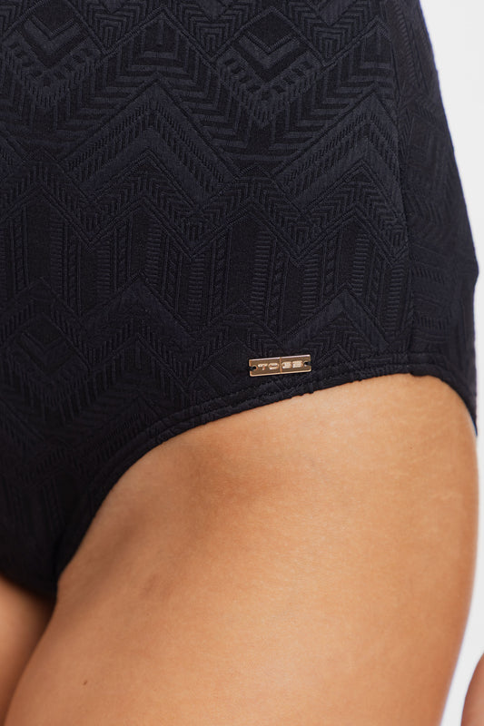 Close-up of a person wearing black textured swimwear bottoms with a geometric pattern and a small gold-tone brand tag near the hem.
