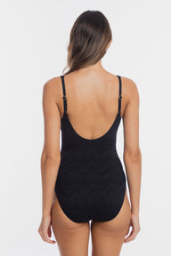 Rear view of a woman wearing a black one-piece swimsuit with thin shoulder straps and a deep open back, photographed against a plain light gray studio background.