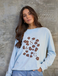 Woman wearing a light blue sweatshirt with floral and monogram design against a textured wall.