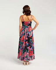 A woman viewed from the back wearing a sleeveless maxi dress with thin straps, a fitted bodice, and a full skirt in a vivid floral print over a black base. She is standing against a plain light gray studio background.