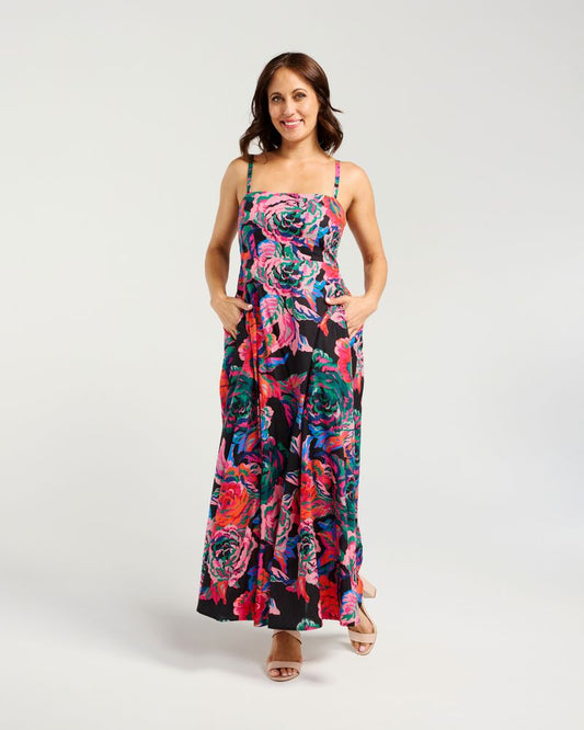 A woman modeling a sleeveless maxi dress with a vibrant floral print in pink, red, blue, and green on a black background, set against a plain light gray studio backdrop.