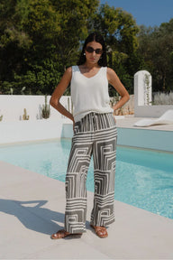 Woman in a white top and patterned pants standing by a pool with greenery in the background