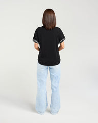 A woman viewed from behind wearing a black short-sleeve top with white striped trim on the sleeve cuffs and light blue wide-leg jeans, against a plain light gray studio background.