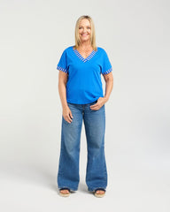 A woman modeling a bright blue short-sleeve top with white and blue striped trim at the V-neck and sleeve cuffs, paired with wide-leg blue jeans, against a light gray studio background.