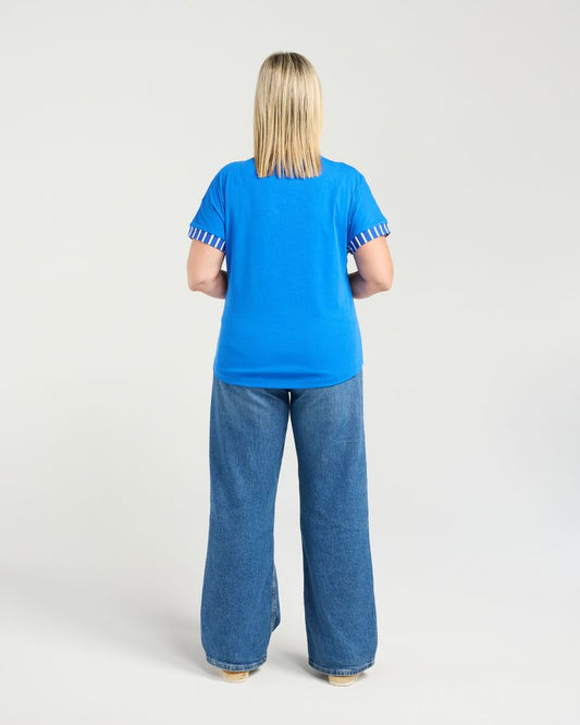 A woman viewed from behind wearing a bright blue short-sleeve t-shirt with white and blue striped ruffle trim on the sleeves, paired with blue jeans, against a light gray studio background.