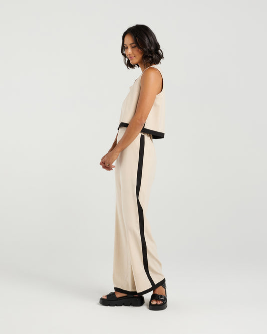 Woman wearing a beige sleeveless top and pants with black trim on a white background