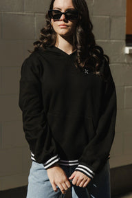 Person wearing a black hoodie with white stripes on the sleeves against a gray wall.