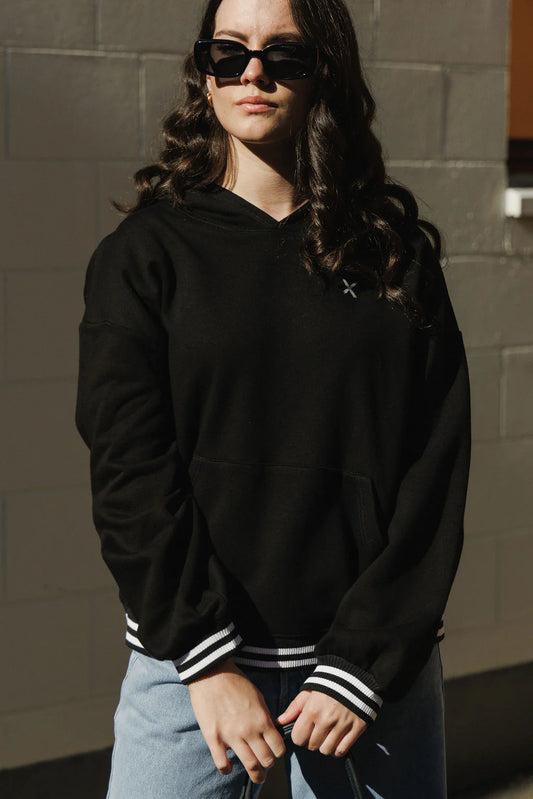 Person wearing a black hoodie with white stripes on the sleeves against a gray wall.