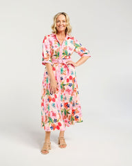 A woman modeling a pink midi shirt dress with a colorful tropical floral print, featuring a collar, three-quarter sleeves, and a matching waist tie, set against a plain light gray studio background.