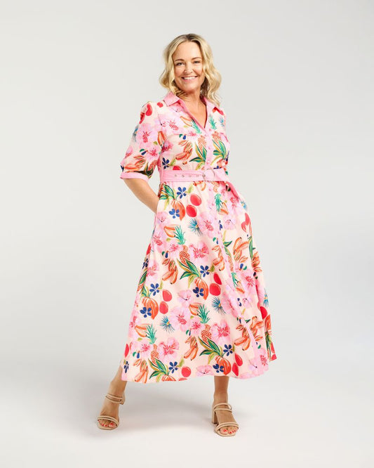 A woman modeling a pink midi dress with a colorful tropical floral and fruit print, featuring short sleeves, a collared neckline, and a matching waist tie, set against a plain light gray studio background.