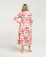 A woman viewed from the back wearing a pink midi wrap dress with a colorful tropical floral print, paired with beige strappy sandals, against a light gray studio background.
