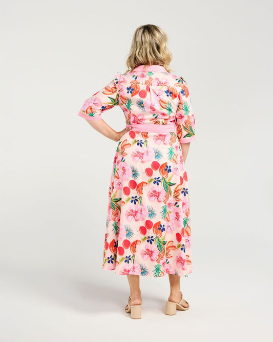 A woman viewed from the back wearing a pink midi wrap dress with a colorful tropical floral print, paired with beige strappy sandals, against a light gray studio background.