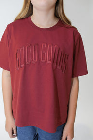 Person wearing a maroon t-shirt with 'GOOD GOODS' text on a plain background
