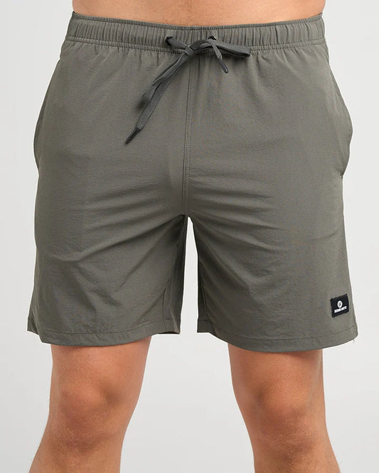 A person wearing olive green drawstring shorts against a light gray studio background. The shorts have an elastic waistband, side pockets, and a small black logo patch near the lower right hem.