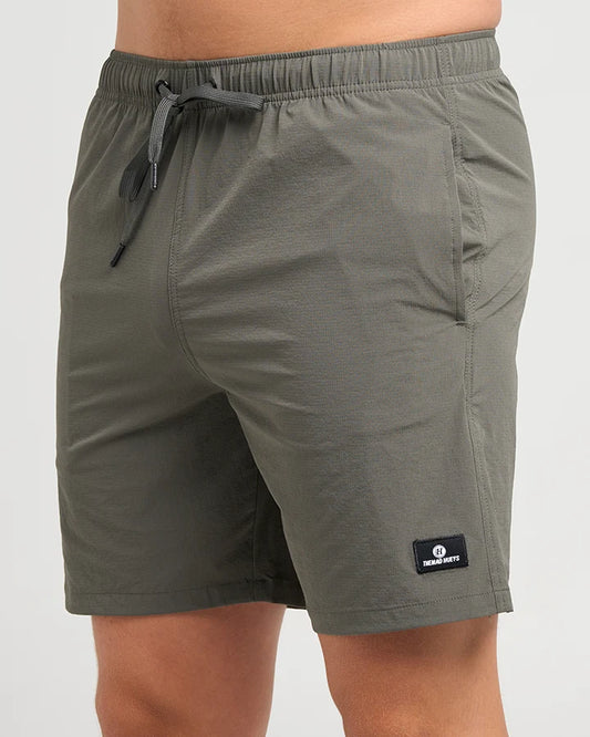 A close-up of a person wearing olive green drawstring shorts, showing the side and front pockets of the shorts. A small black brand patch is visible on the lower left leg.