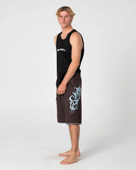 A man stands in profile wearing a black sleeveless tank top and dark brown pinstriped board shorts with a large light blue graphic on the left leg, photographed against a light gray studio background.