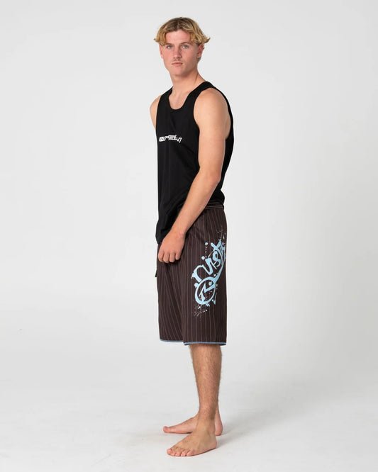 A man stands in profile wearing a black sleeveless tank top and dark brown pinstriped board shorts with a large light blue graphic on the left leg, photographed against a light gray studio background.