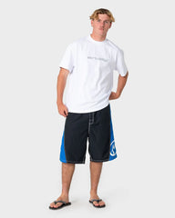 A man modeling a white short-sleeve t-shirt with a gray logo on the chest and dark navy board shorts with bright blue side panels and a white logo, paired with black flip-flops, against a light gray background.