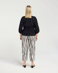 A woman viewed from the back wearing a black pleated long-sleeve top and black-and-white vertically patterned pants, standing against a light gray studio background.