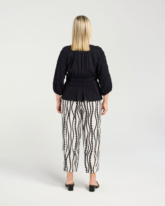 A woman viewed from the back wearing a black pleated long-sleeve top and black-and-white vertically patterned pants, standing against a light gray studio background.