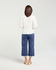 A woman viewed from behind wearing a white pleated long-sleeve top and blue cropped pants, standing against a light gray studio background.