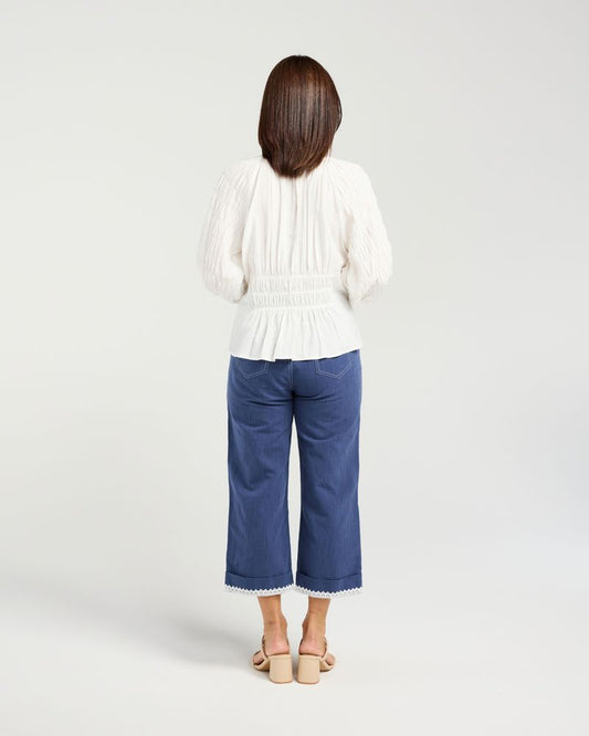 A woman viewed from behind wearing a white pleated long-sleeve top and blue cropped pants, standing against a light gray studio background.