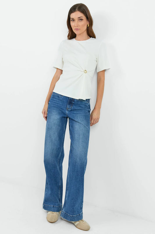 A woman modeling a white short-sleeve top with a twisted front detail and a gold button, paired with wide-leg blue jeans and beige shoes, against a plain white studio background.