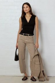 A woman stands against a white brick wall wearing a black sleeveless wrap top, beige wide-leg cropped pants, a black belt, and black strappy sandals. She is holding a black handbag and a beige jacket draped over her arm.