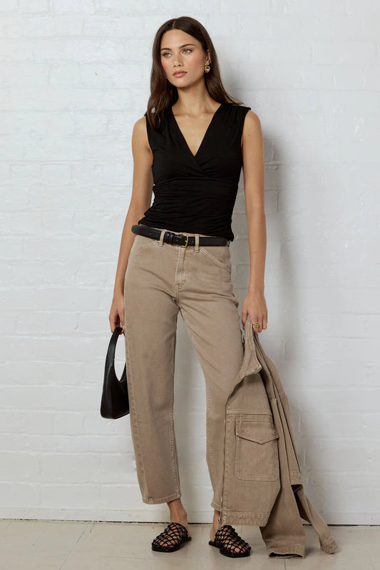 A woman stands against a white brick wall wearing a black sleeveless wrap top, beige wide-leg cropped pants, a black belt, and black strappy sandals. She is holding a black handbag and a beige jacket draped over her arm.
