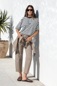 A woman stands outdoors against a white wall wearing a black and white striped short-sleeve top, beige cropped trousers, and leopard-print mule flats, with a beige jacket tied around her waist.