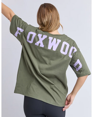 A woman shown from the back wearing an olive green short-sleeve T-shirt with large light purple 'FOXWOOD' lettering across the upper back, paired with dark leggings, against a light gray studio background.