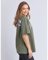 A woman wearing an oversized olive green t-shirt with light purple graphic lettering on the back, photographed against a light gray studio background.