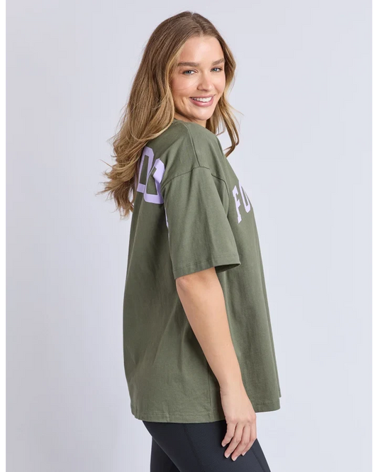 A woman wearing an oversized olive green t-shirt with light purple graphic lettering on the back, photographed against a light gray studio background.