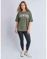 A woman modeling an oversized olive green t-shirt with 'FOXWOOD' printed in white across the chest, paired with dark leggings and white sneakers, against a light gray studio background.