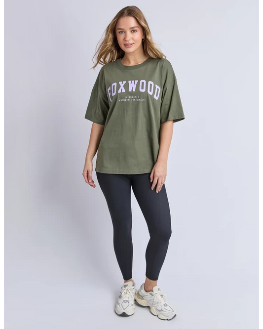 A woman modeling an oversized olive green t-shirt with 'FOXWOOD' printed in white across the chest, paired with dark leggings and white sneakers, against a light gray studio background.