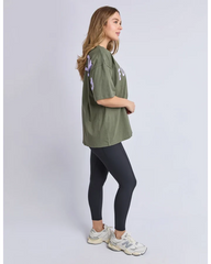 A woman modeling an oversized olive green t-shirt with light purple and white graphic prints on the sleeves and chest, paired with black leggings and white sneakers, shown from a side profile against a light gray studio background.