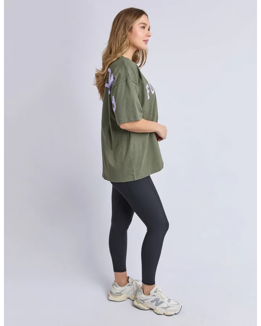 A woman modeling an oversized olive green t-shirt with light purple and white graphic prints on the sleeves and chest, paired with black leggings and white sneakers, shown from a side profile against a light gray studio background.