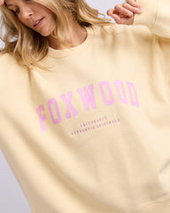 Yellow sweatshirt with pink 'FOXWOOD' text worn by a person on a light gray background