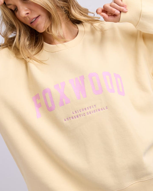 Yellow sweatshirt with pink 'FOXWOOD' text worn by a person on a light gray background