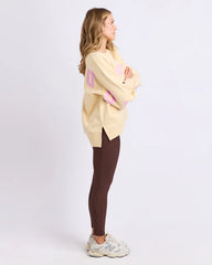 Woman wearing a light yellow oversized sweater with pink letters and dark brown leggings on a white background