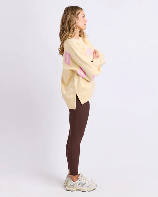 Woman wearing a light yellow oversized sweater with pink letters and dark brown leggings on a white background